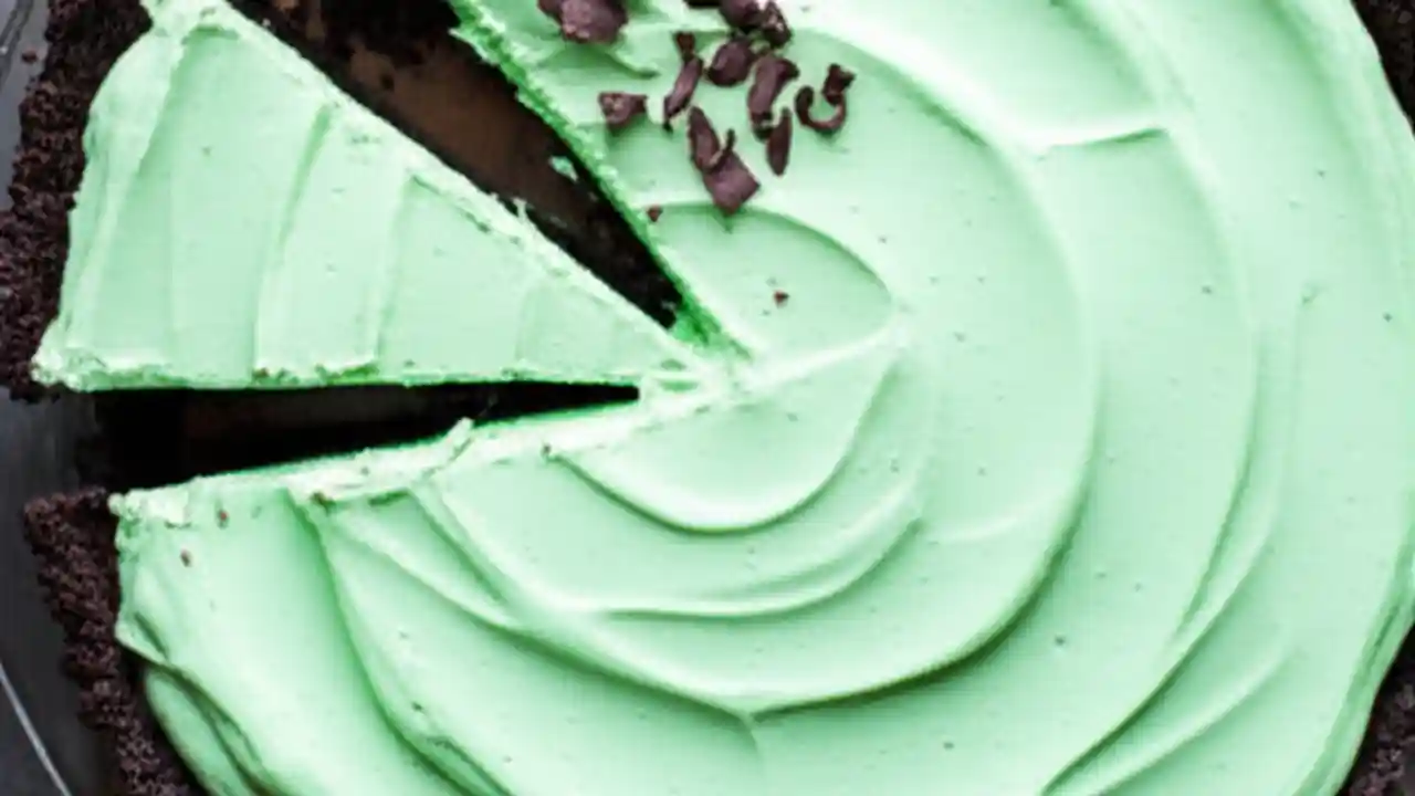 A perfect slice of vibrant green Grasshopper Pie with a chocolate crust, garnished with mint and chocolate shavings, on a wooden board.